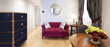 Een kamer met een rode bank, een houten tafel met een fruitschaal, een blauwe dressoir en een ronde spiegel aan de muur.