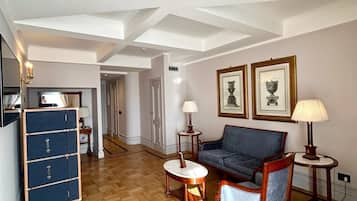 A spacious living area with a wooden parquet floor, a blue sofa, a small round table, and a blue cabinet. There are framed pictures on the wall and a staircase in the background.