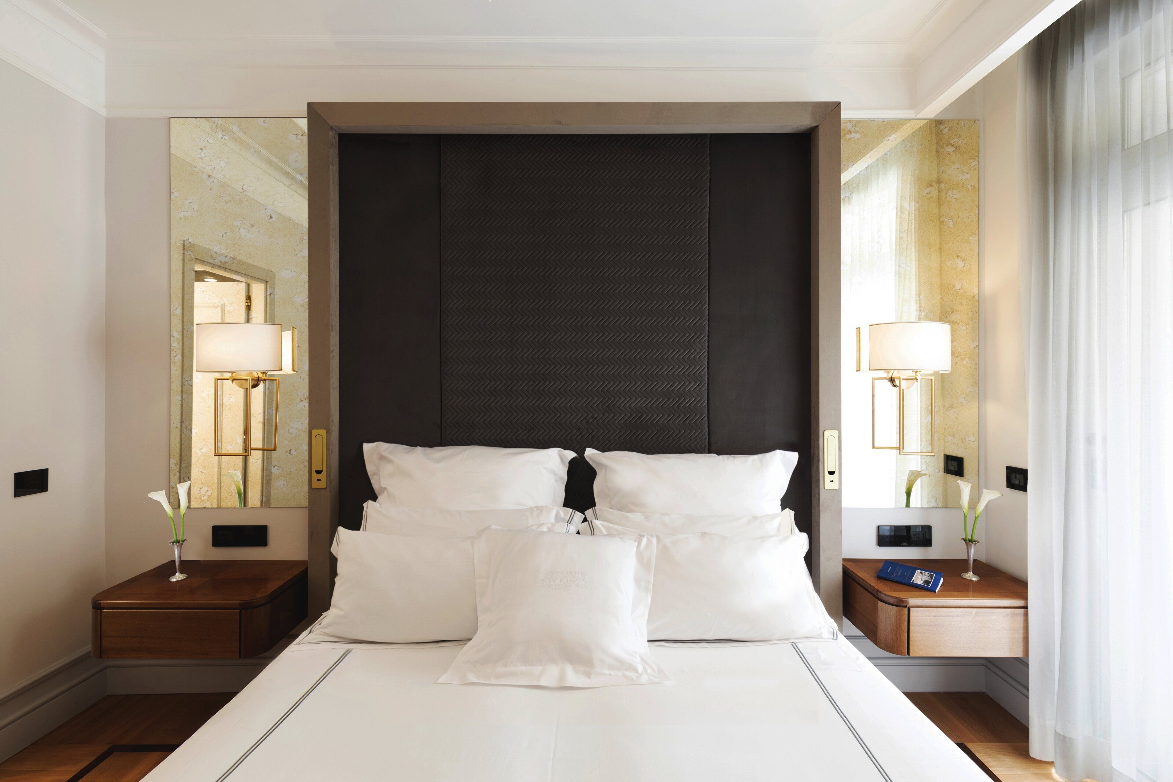 A hotel room with a large bed, two bedside tables with lamps, a dark headboard, and a mirror on the wall.