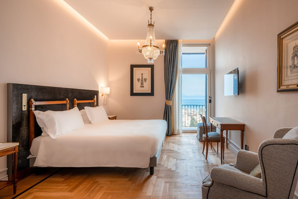 A hotel room with a large bed, a desk, a chair, a chandelier, and a framed picture on the wall.