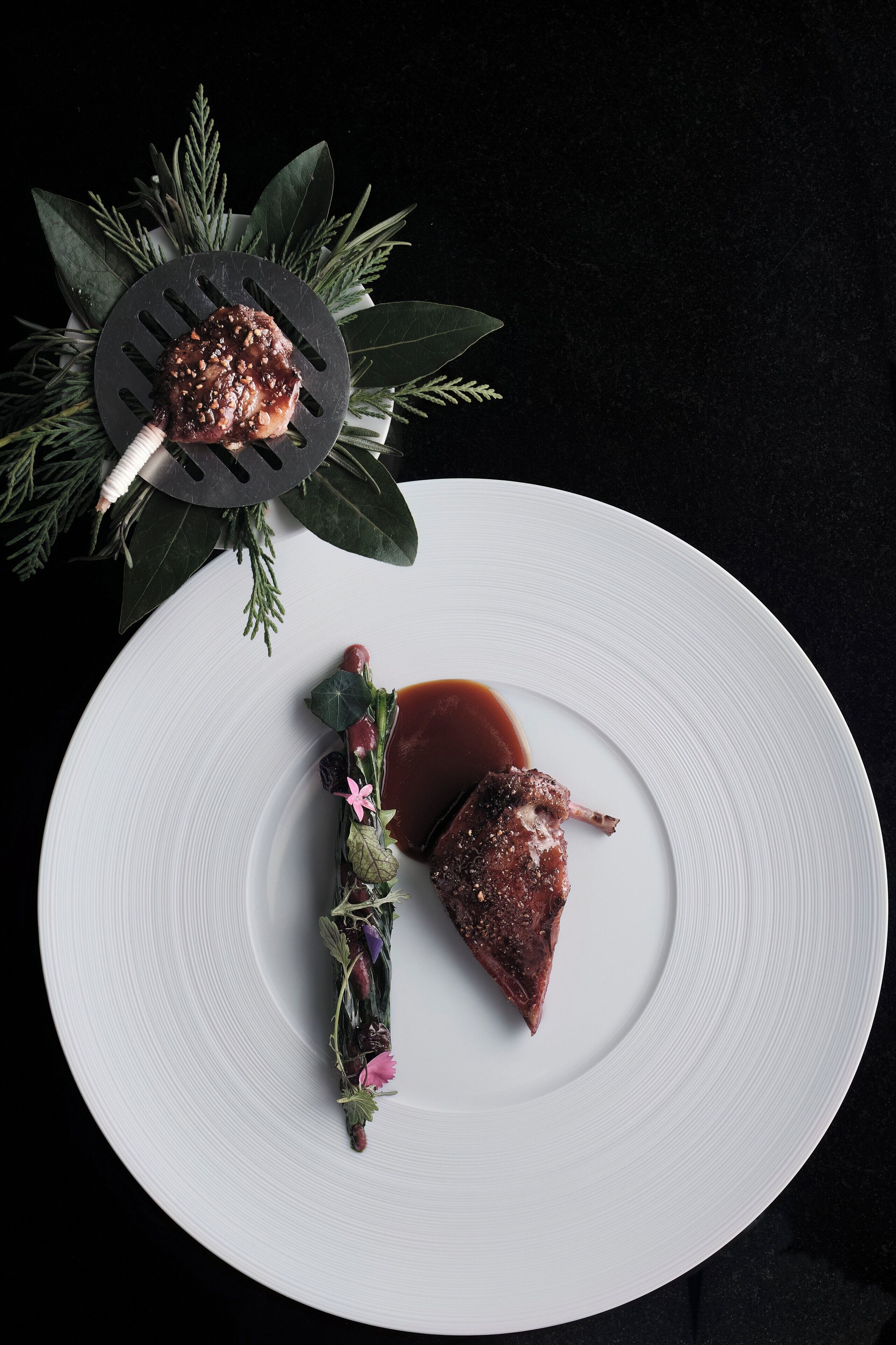 A white plate with a piece of meat and sauce, garnished with herbs and flowers, on a dark background.