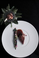 A white plate with a piece of meat and sauce, garnished with herbs and flowers, on a dark background.