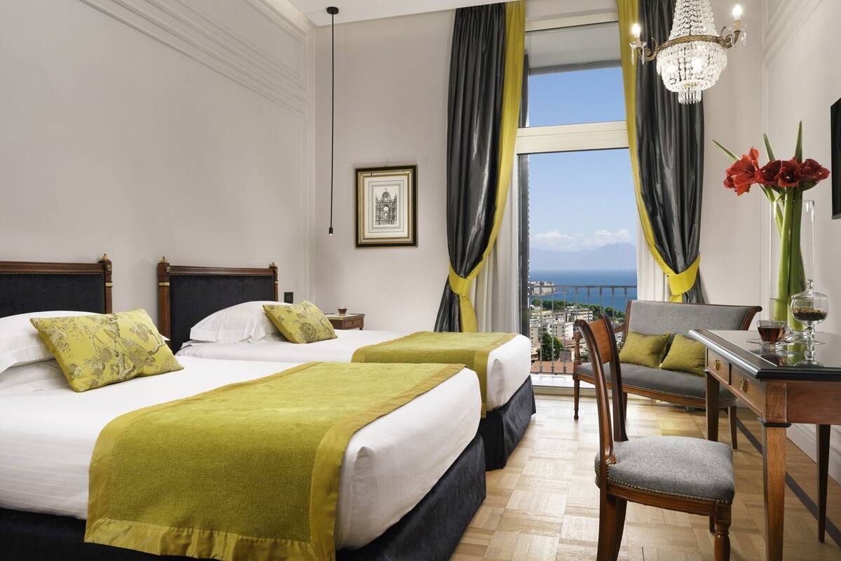 A hotel room with two beds, a desk, a chair, a chandelier, and a view of the sea.