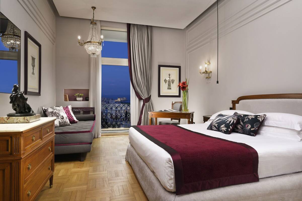 A hotel room with a bed, a desk, a dresser, a chandelier, and a view of the city at night.