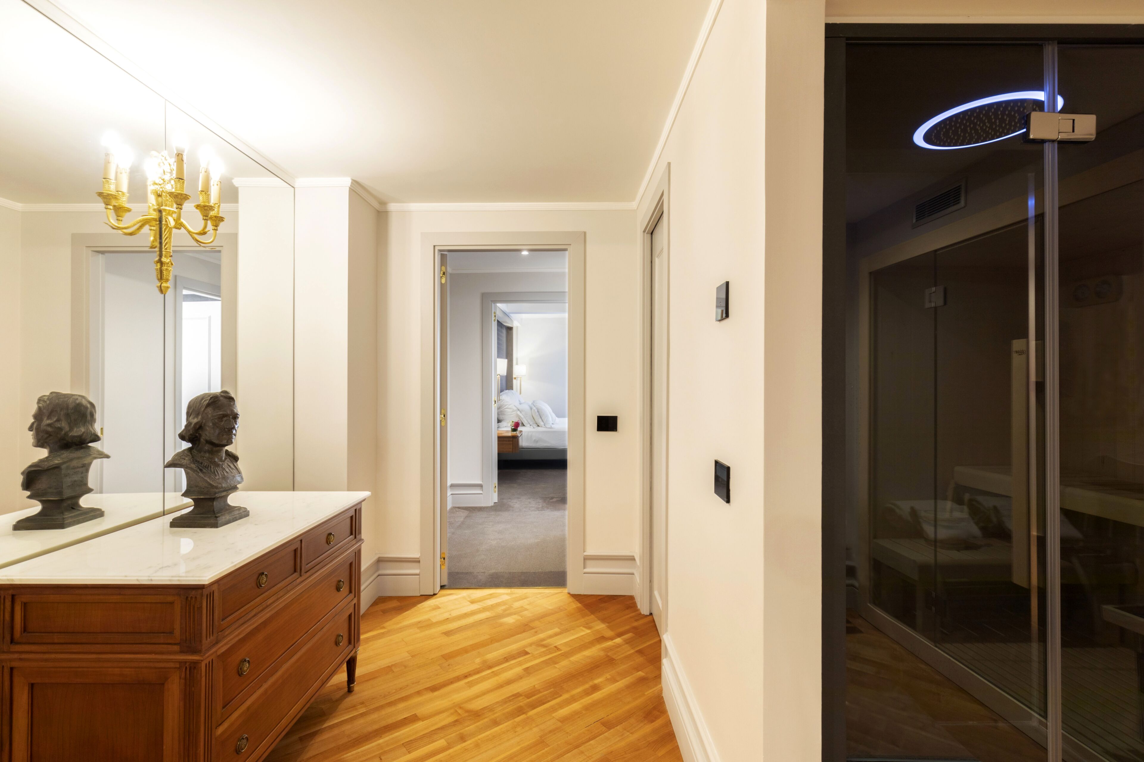 A hallway with a wooden floor, a marble countertop with bust sculptures, a chandelier, and a glass door with a blue light.
