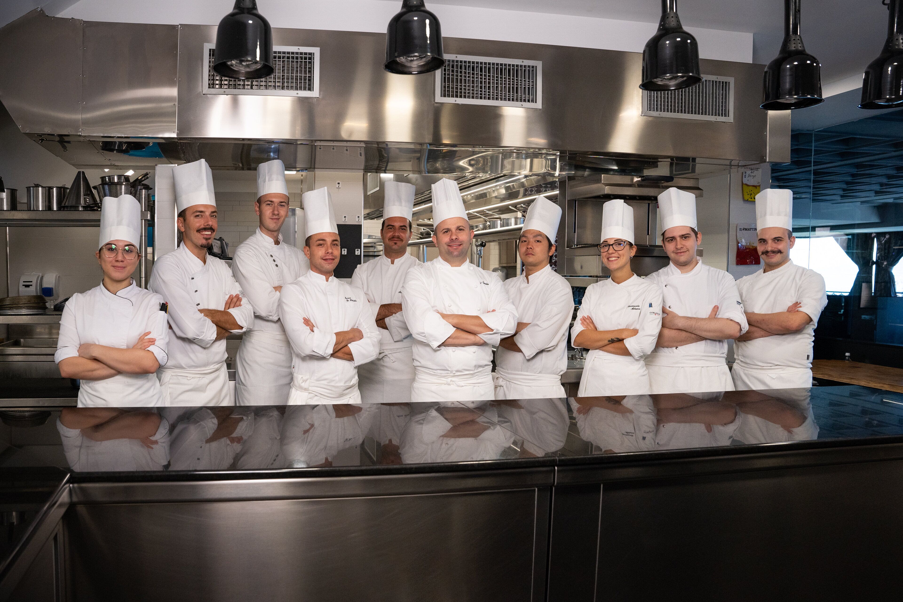 A group of chefs standing in a kitchen with stainless steel appliances and overhead lighting.