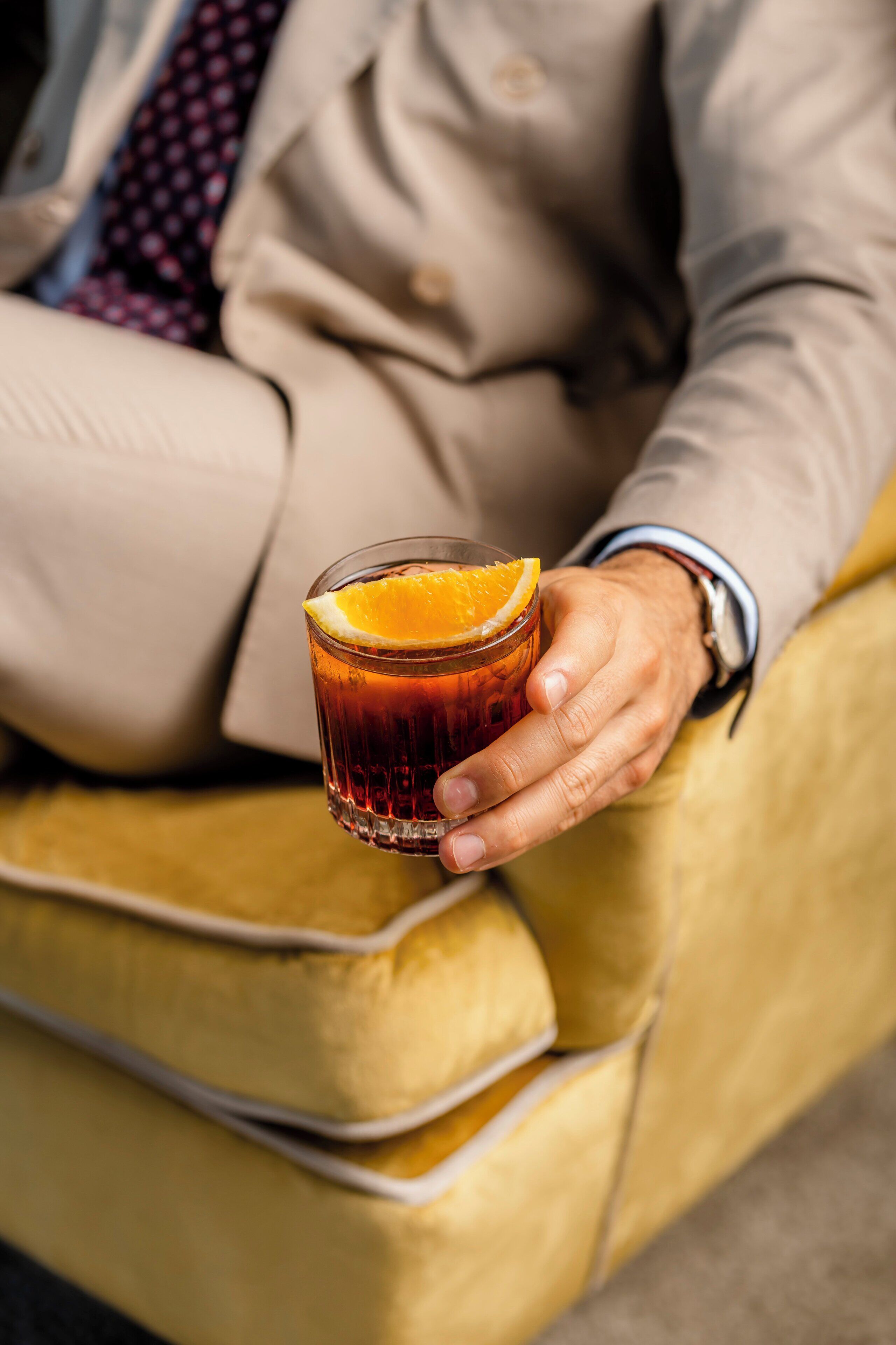 A person holding a glass of amber liquid with an orange slice garnish, seated on a yellow upholstered chair.