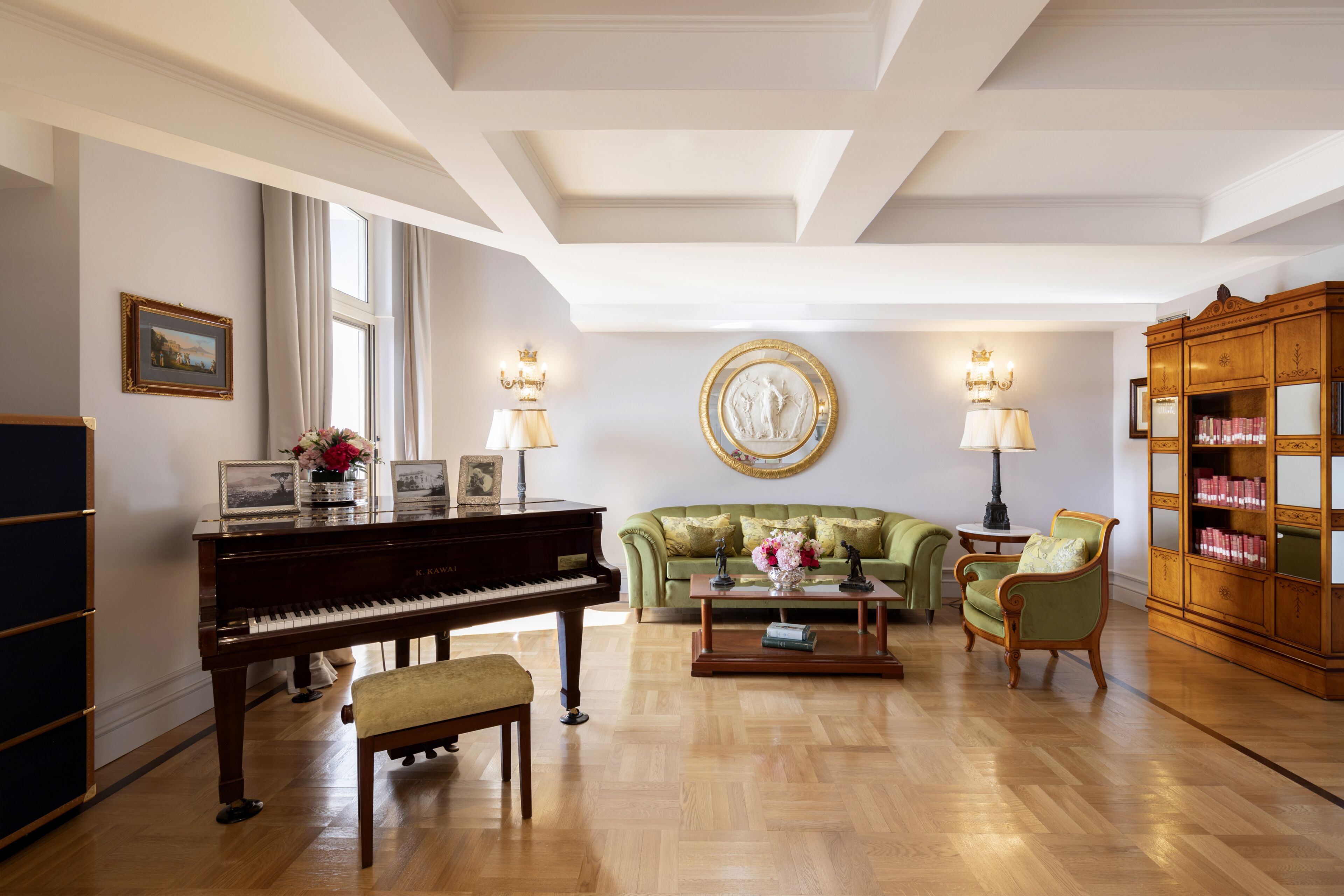 Een ruime woonkamer met een vleugelpiano, een groene bank, een salontafel en een houten boekenkast.
