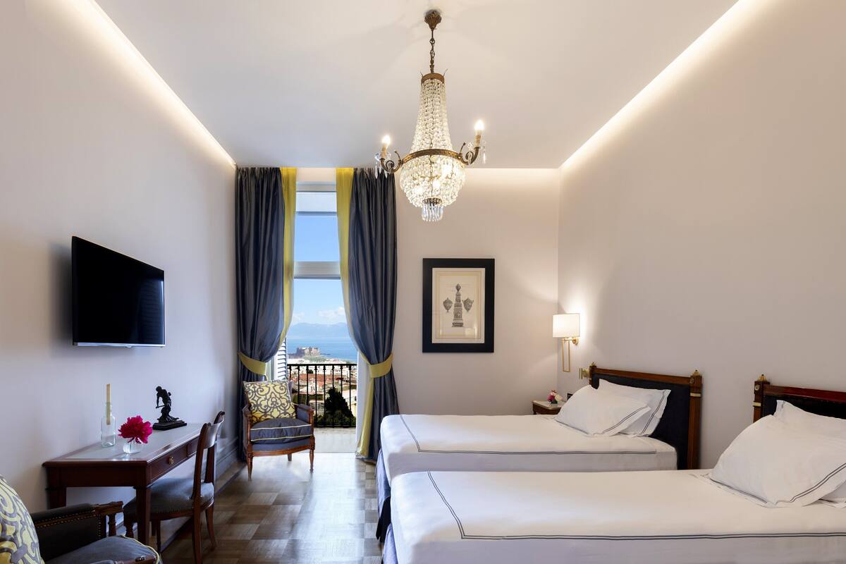 A hotel room with two beds, a desk, a chair, a TV, a chandelier, and a window with curtains.