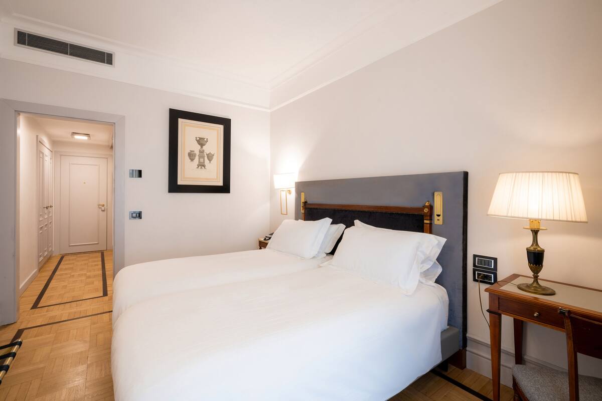 A hotel room with a double bed, a bedside table with a lamp, a framed picture on the wall, and a door leading to a hallway.