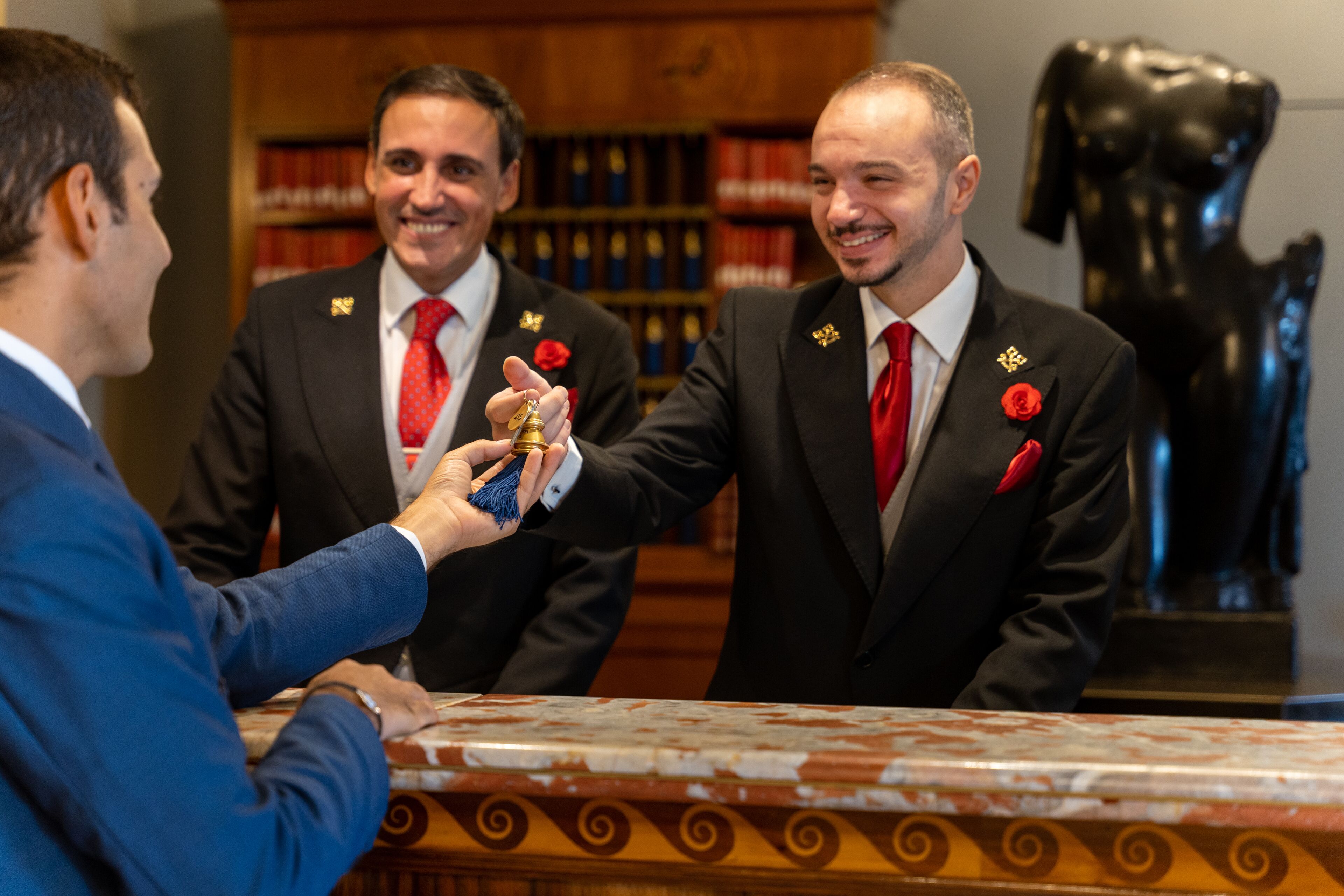 Three men in suits with red ties and lapel pins, standing in a room with a marble table and bookshelves in the background.