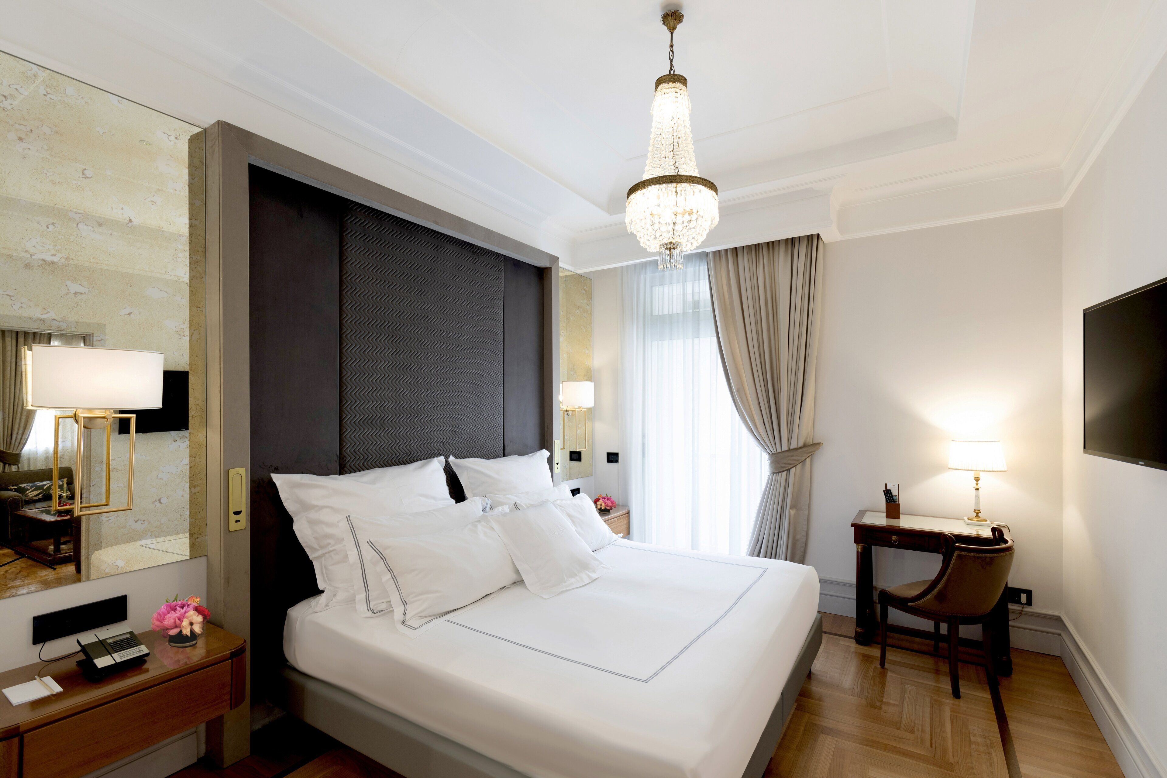 A hotel room with a large bed, a desk, a chair, a TV, and a chandelier.