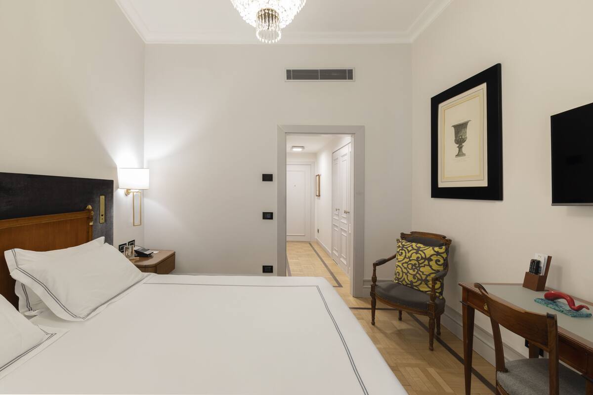A hotel room with a large bed, a bedside table with a lamp, a chair, a desk with a chair, and a framed picture on the wall.