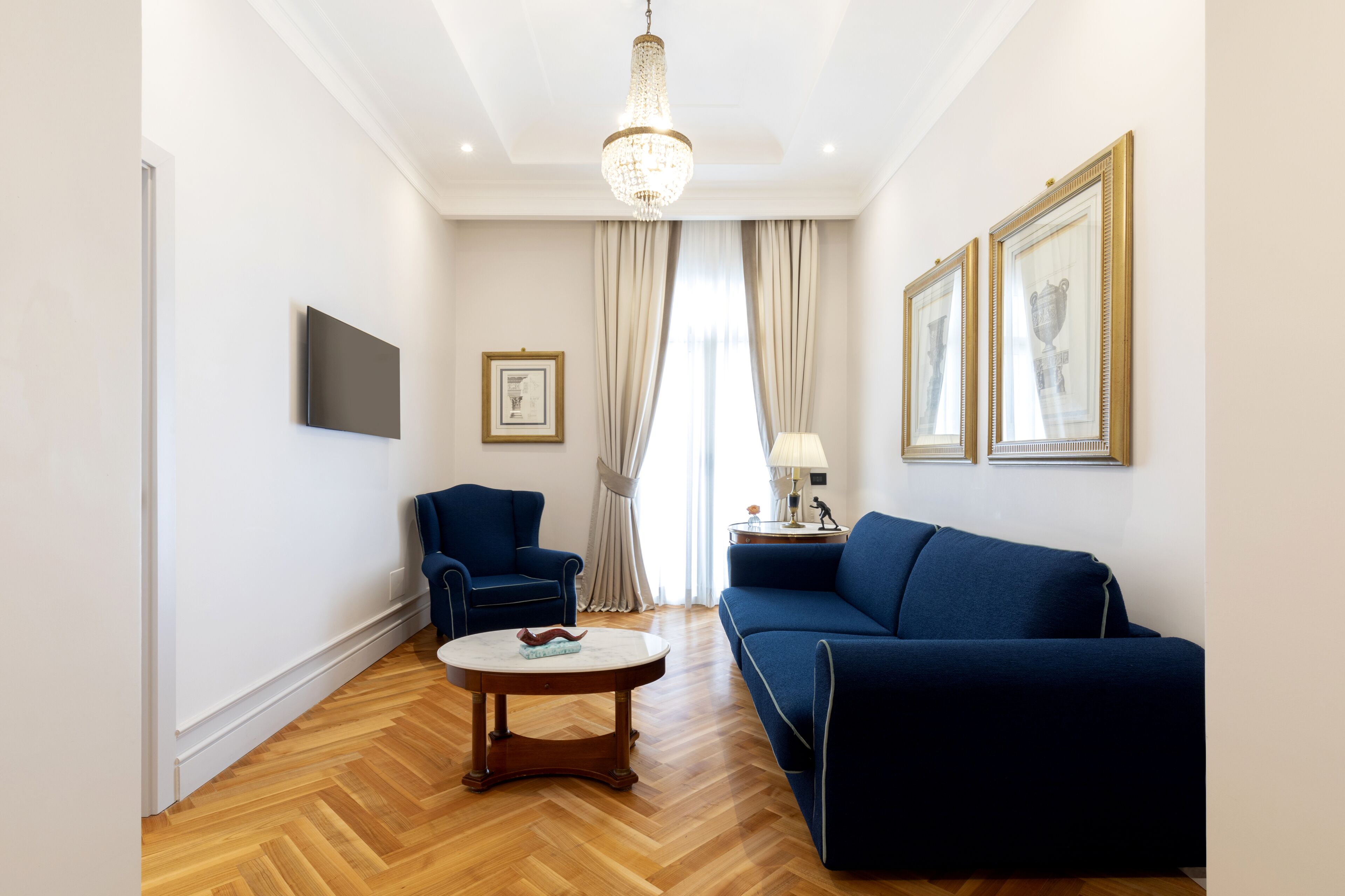 A modern living room with a blue sofa, a blue armchair, a round coffee table, a chandelier, and framed artwork on the walls.