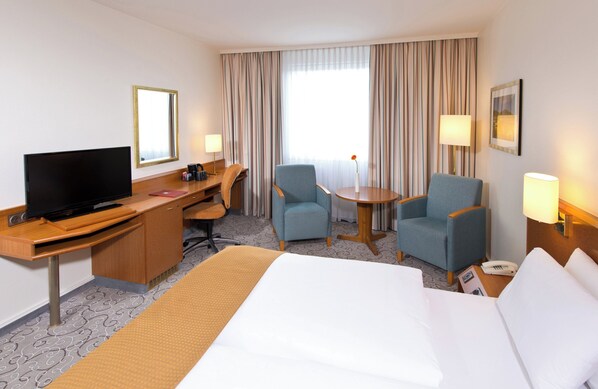 Una habitación de hotel con una cama, un escritorio con televisor, dos sillones, una mesita y una lámpara.
