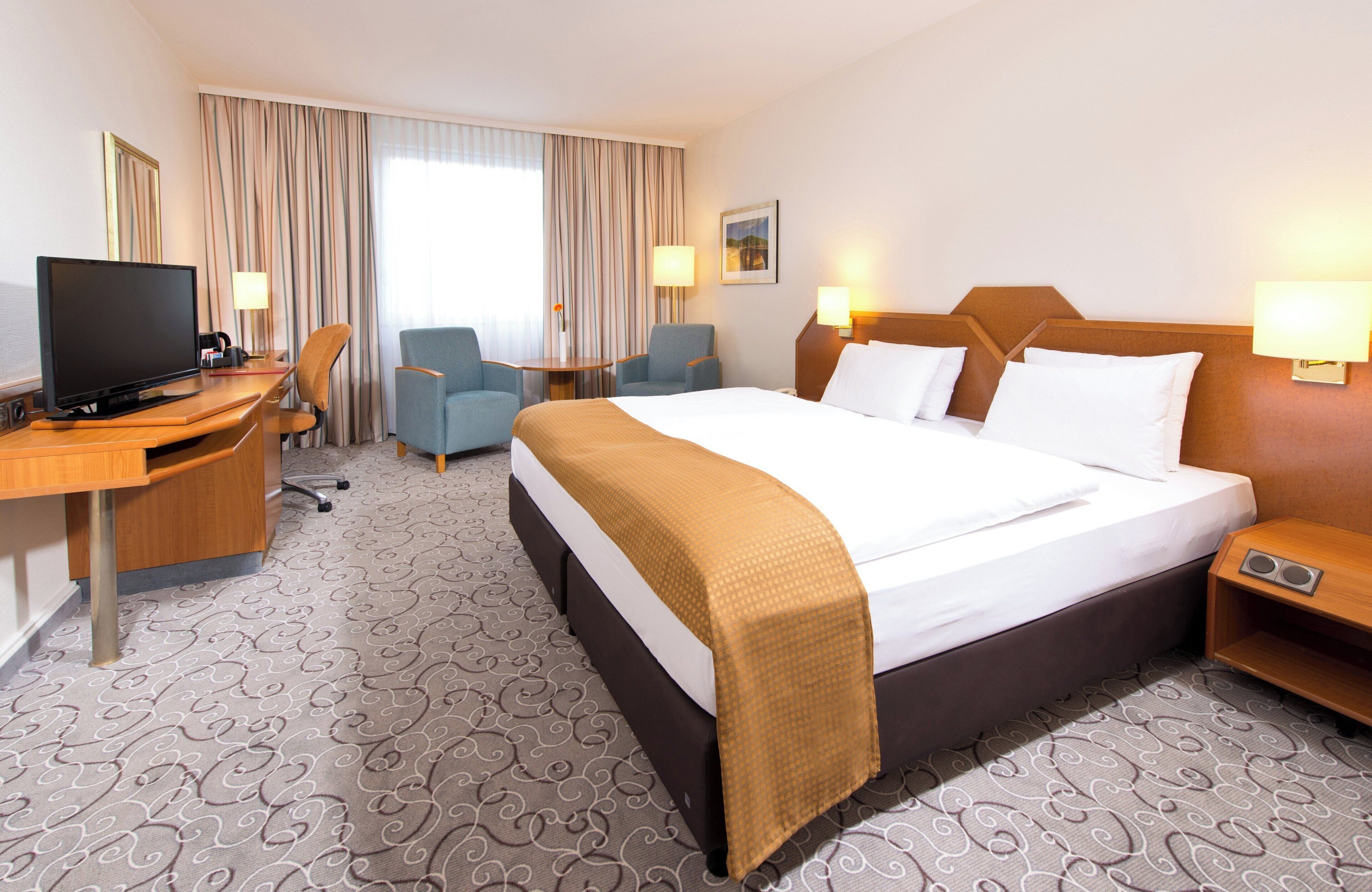 A hotel room with a large bed, a desk with a television, two armchairs, a small table, and a window with curtains.