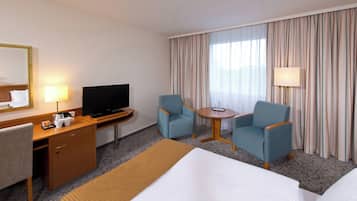 A hotel room with a bed, a desk with a television, two armchairs, a small table, and a large window with curtains.