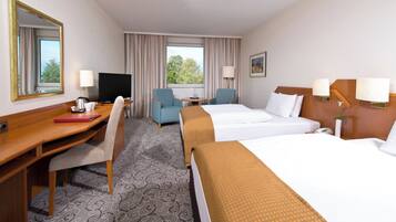 A hotel room with a desk, two beds, a TV, and a view of trees through the window.