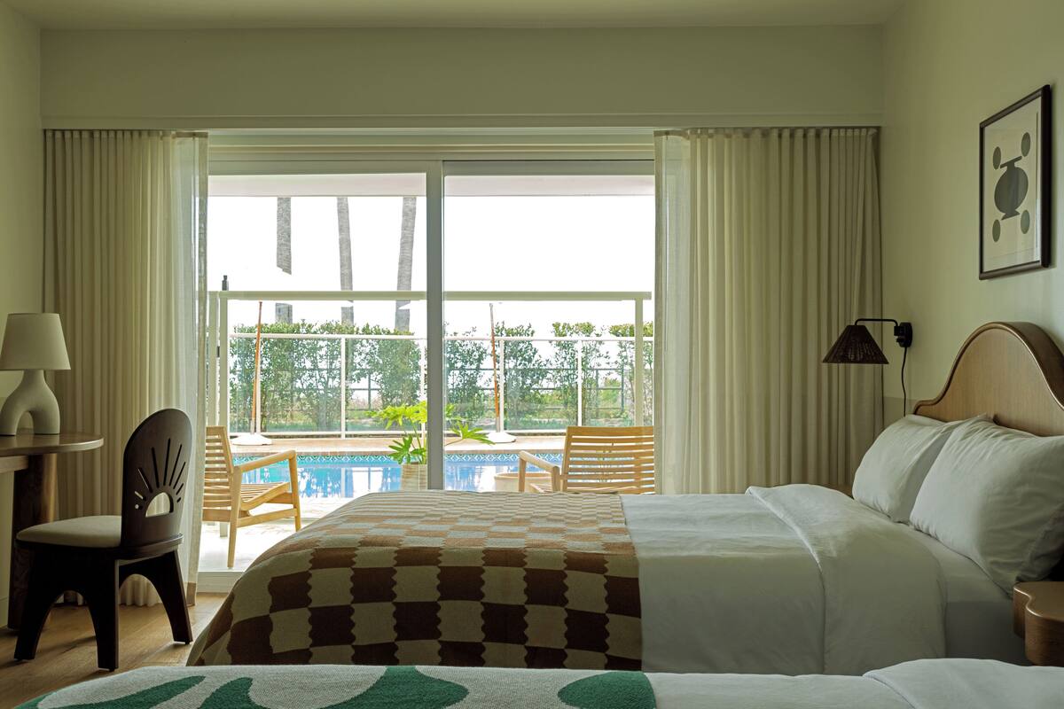 A hotel room with a bed, a chair, a table, and a view of a pool and greenery outside.
