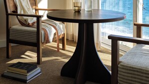 A room with a round table, two chairs, a bookshelf, and a view of the ocean through large windows.