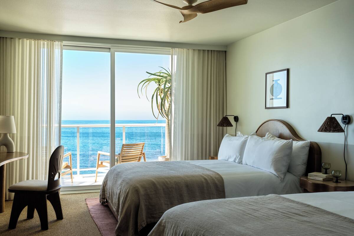A hotel room with two beds, a desk, a chair, a plant, and a view of the ocean.