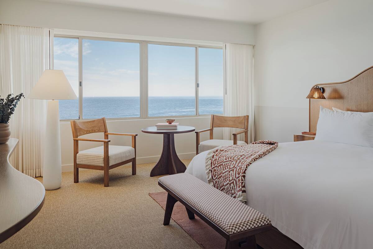 A hotel room with a large window, a bed, two chairs, a small table, a bench, and a lamp.