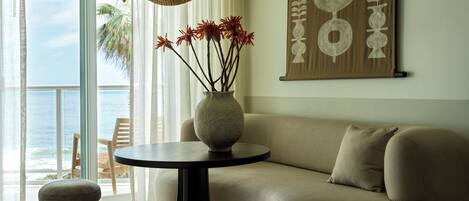 A living room with a sofa, a round table with a vase of flowers, a hanging light fixture, and a large window with a view of the ocean.