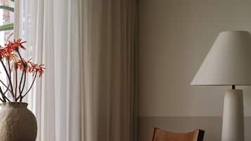 A room with a wooden chair, a table with a vase of red flowers, a lamp, and sheer curtains.