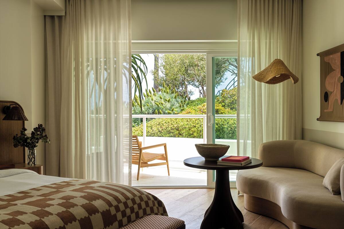 A hotel room with a bed, a sofa, a table with a bowl and books, a lamp, and a view of greenery through a sliding door.