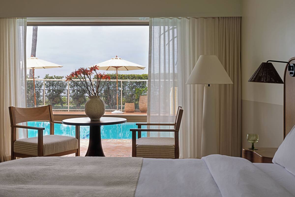 A hotel room with a bed, two chairs, a table with a vase, a lamp, and a view of a pool and greenery.