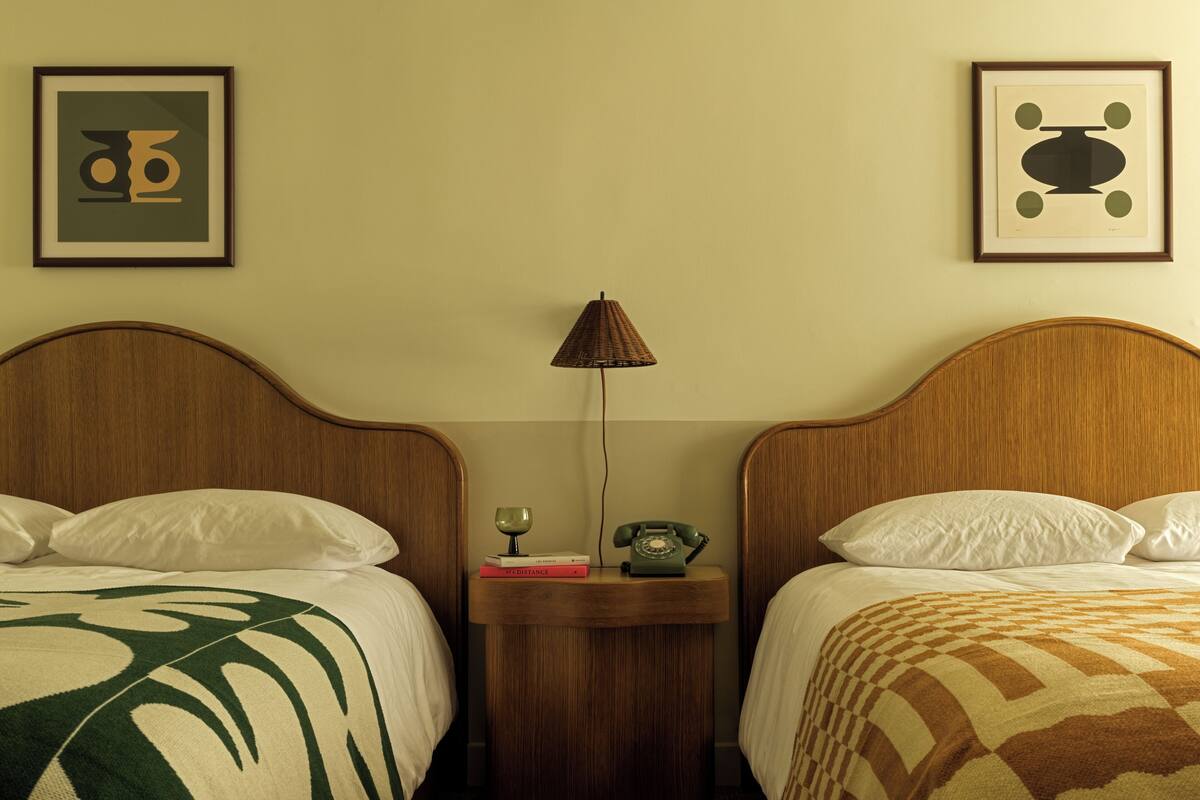 A hotel room with two beds, a nightstand with a lamp, a telephone, and framed artwork on the wall.