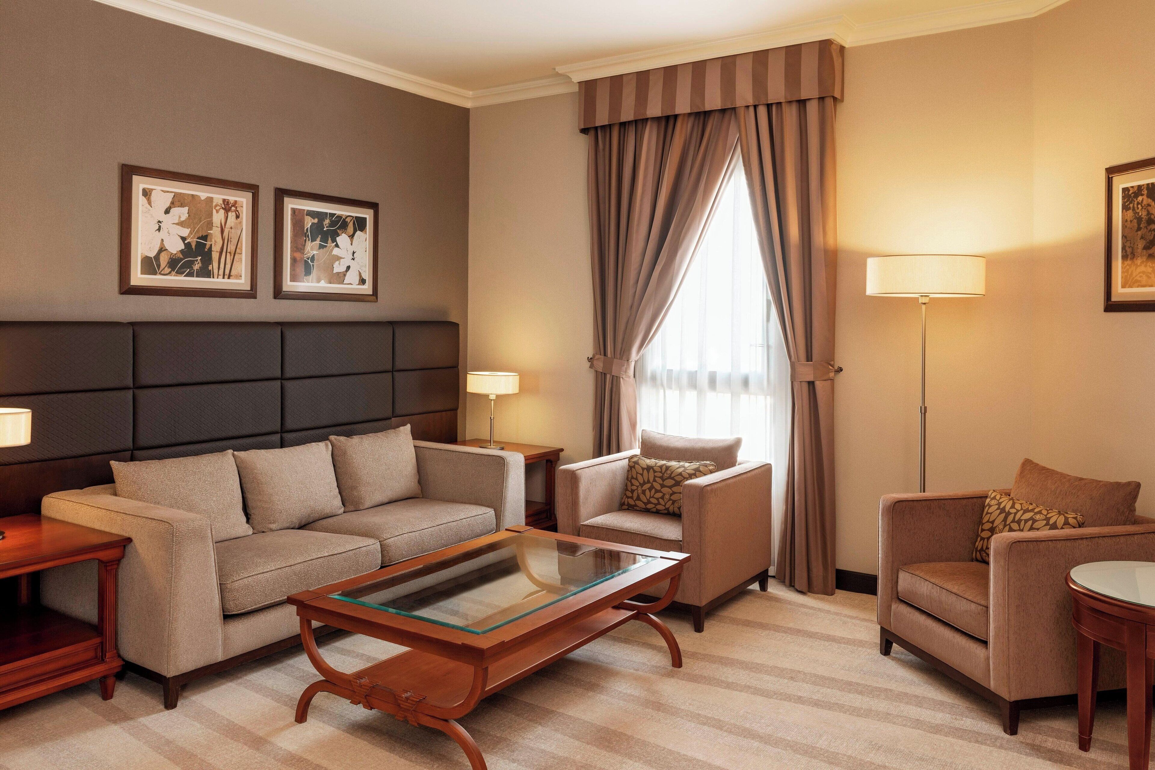A hotel room with a sofa, armchair, and a small table. There are two lamps and a window with curtains.