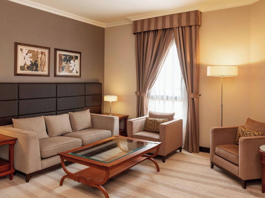 Habitación de hotel con sofá, sillón y una pequeña mesa. Hay dos lámparas y una ventana con cortinas.