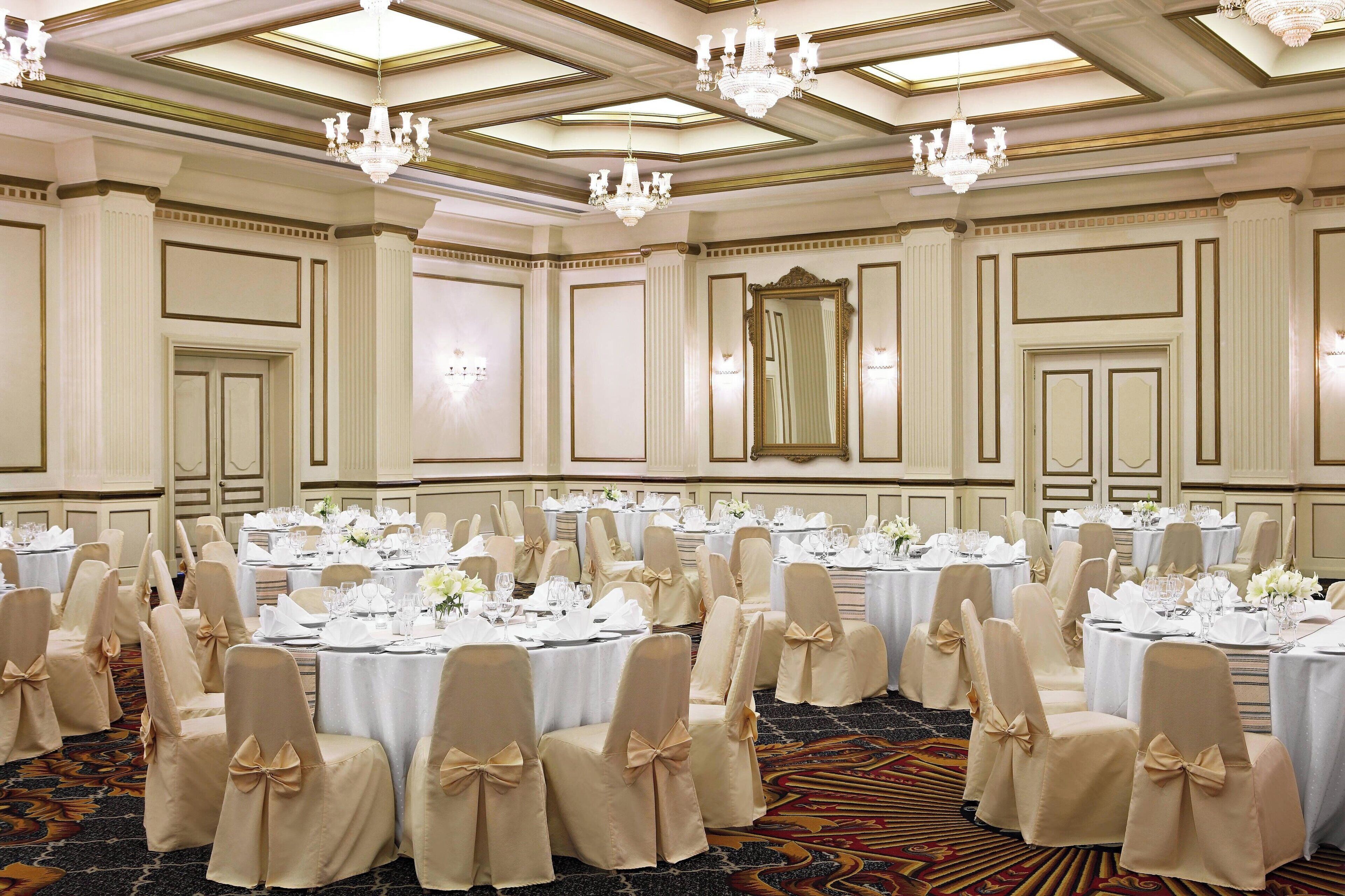 A banquet hall with round tables set for a formal event, featuring cream-colored chairs with bows, chandeliers, and a classic ceiling design.