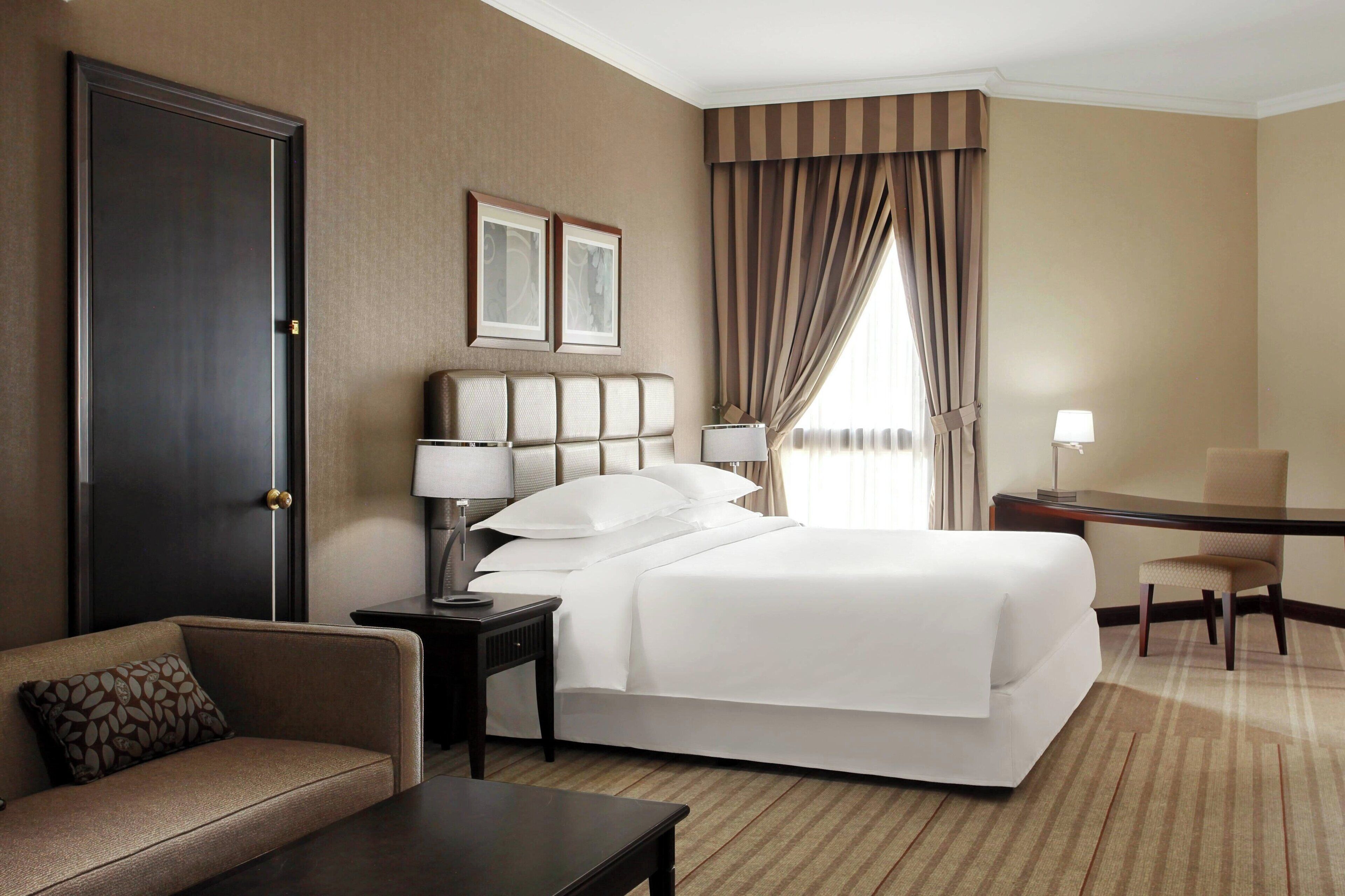A hotel room with a large bed, a desk, a chair, a sofa, and a door.