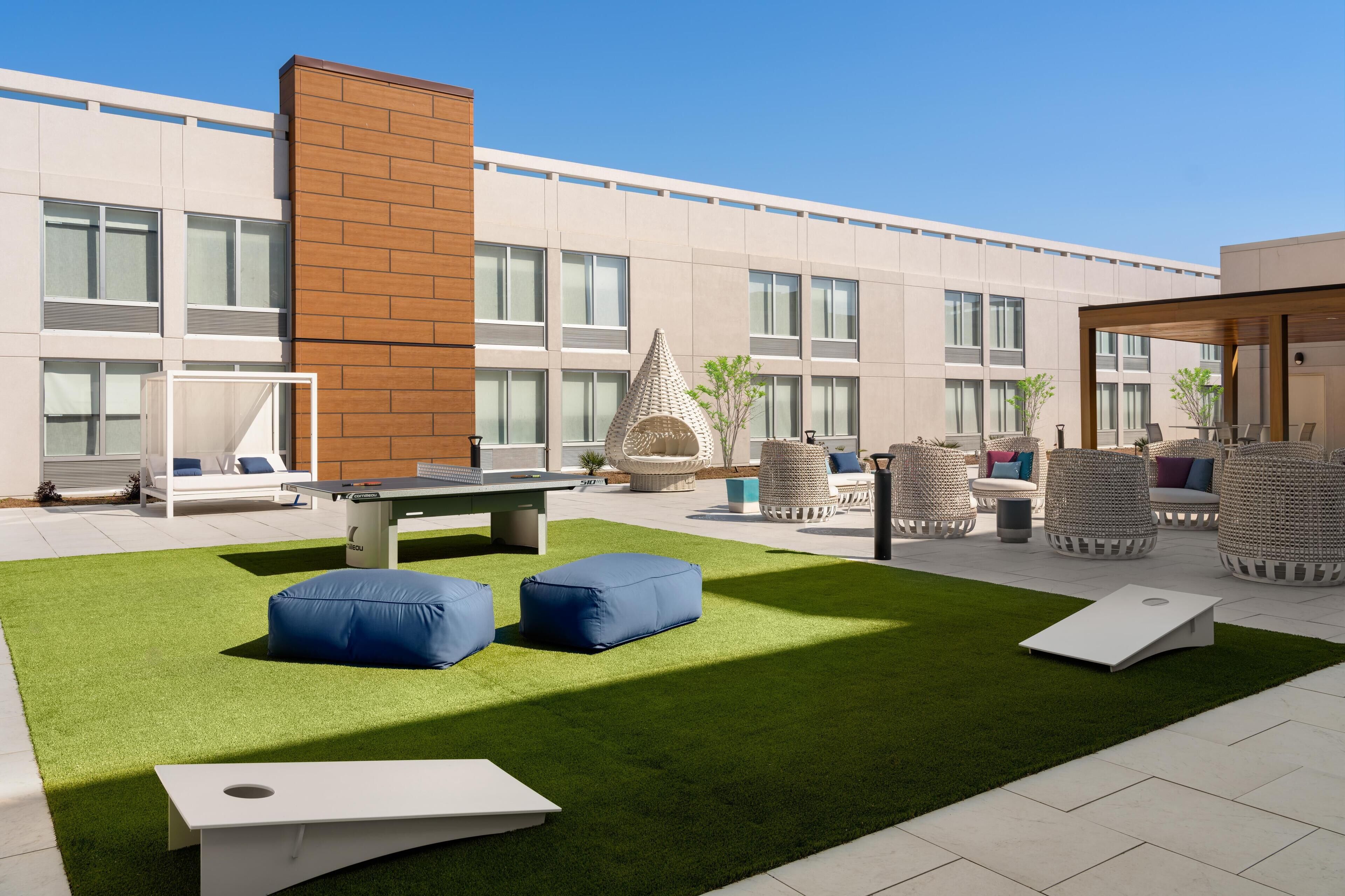 Een terras buiten met een groen gazon, witte tafels, blauwe kussens, rieten stoelen en een modern gebouw op de achtergrond.