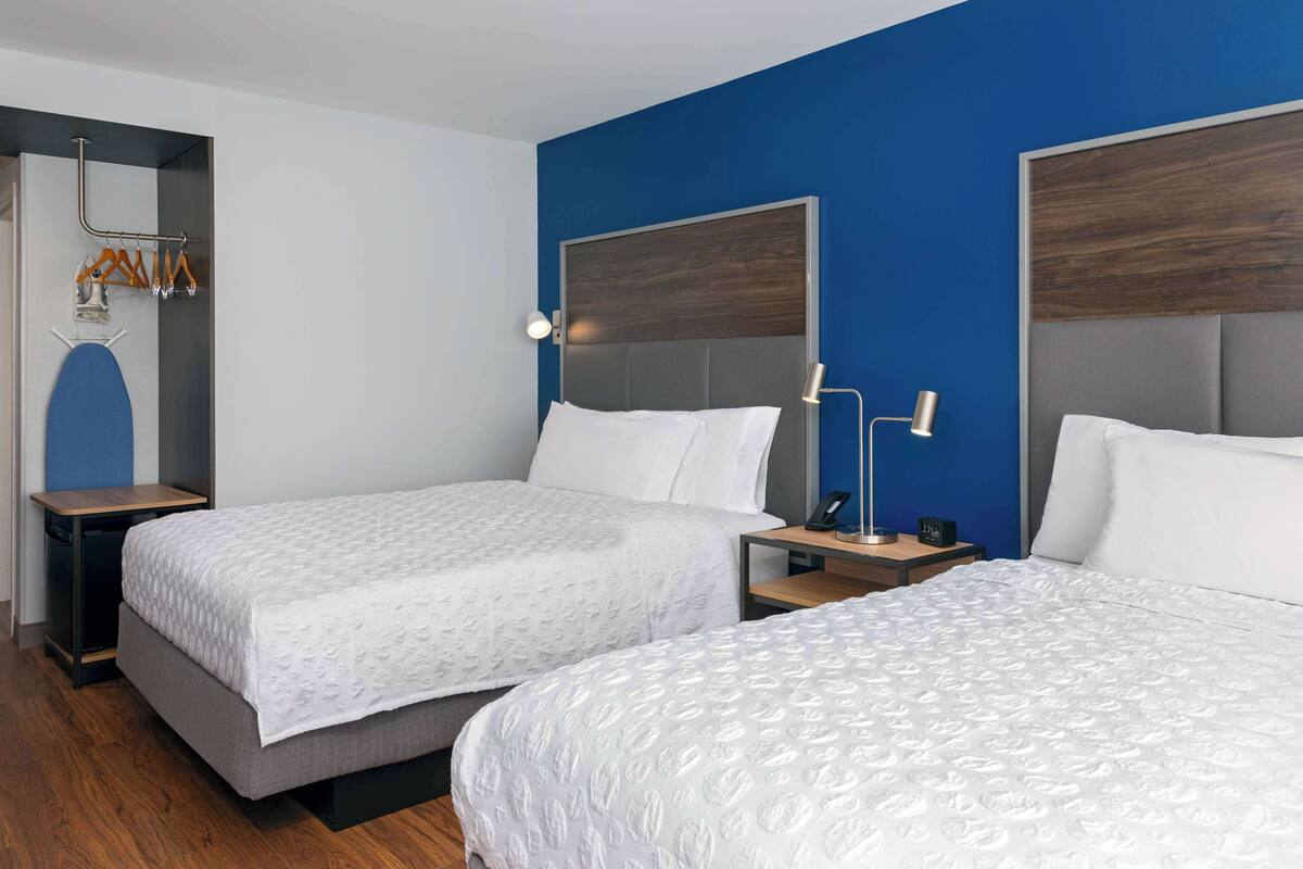 Ein Hotelzimmer mit zwei Betten, Holzboden, einer blauen Akzentwand und einem Kleiderständer mit Kleiderbügeln.