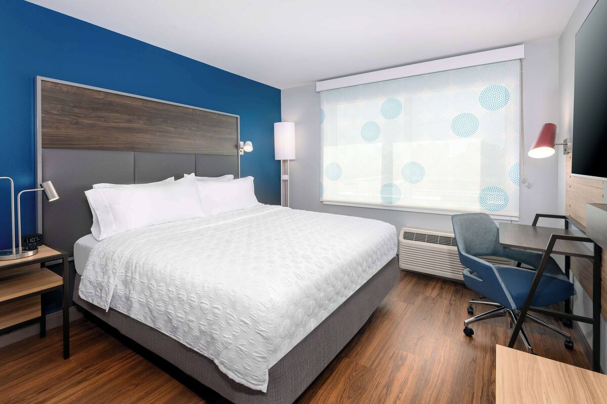 Ein modernes Hotelzimmer mit einem großen Bett, einem Schreibtisch, einem Sessel und einem großen Fenster mit Blick auf die Stadt.