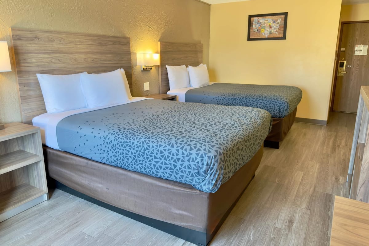 Une chambre d’hôtel avec deux lits, une tête de lit en bois, une table de chevet, une lampe fixée au mur et un tableau encadré.
