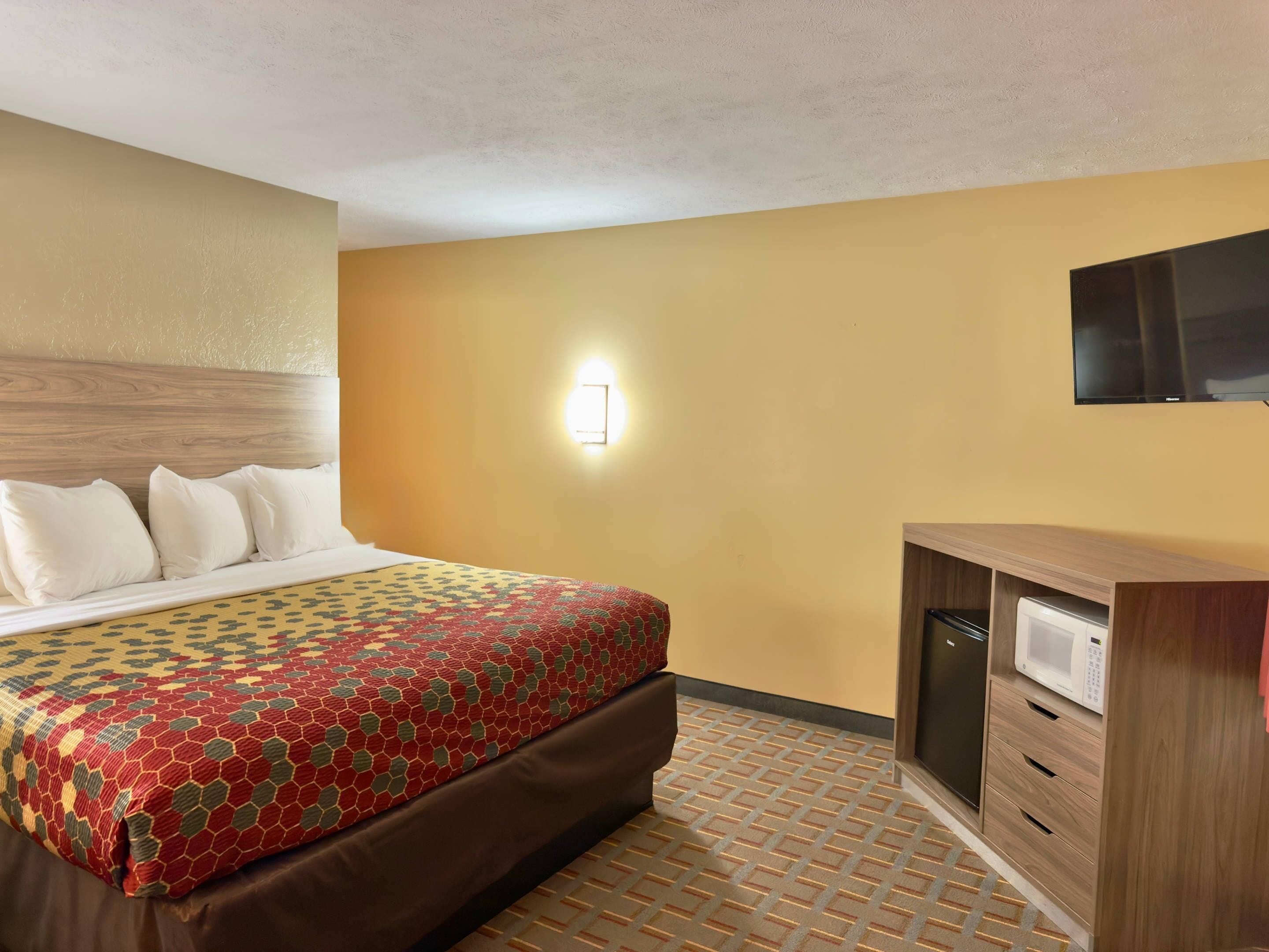 Une chambre d’hôtel comprenant un lit, un téléviseur fixé au mur, une table de chevet avec un micro-ondes et un tapis à motifs.