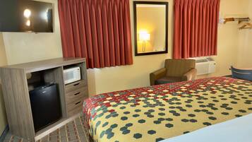 A hotel room with a bed, a desk with a microwave, a chair, a TV, and a window with curtains.