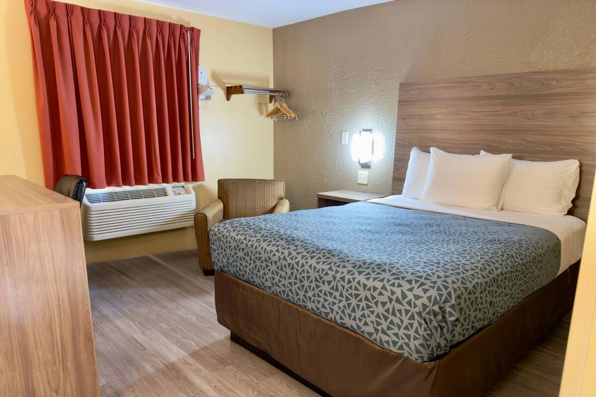 Une chambre d’hôtel avec un lit, une chaise, un bureau, un climatiseur fixé au mur et une fenêtre avec des rideaux rouges.