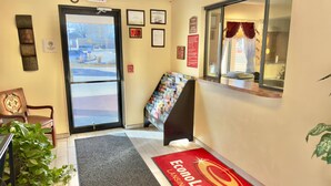A hotel lobby with an exit sign, framed certificates, a fire alarm, a reception desk, a plant, and a red carpet with the hotel name. - Econo Lodge Lansing - Leavenworth (Lansing)