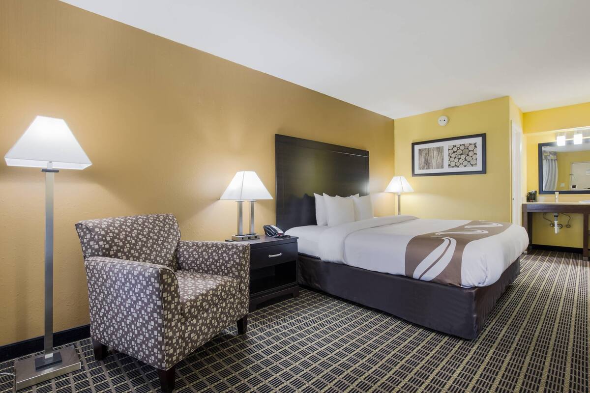 Quarto de hotel com cama, duas luminárias de cabeceira, uma cadeira, mesa de cabeceira, cabeceira, obra de arte na parede e espelho de banheiro.