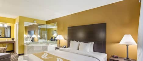 A hotel room with a large bed, a sitting area with a chair, a desk with a lamp, a mirror, and a bathroom with a bathtub and mirror.