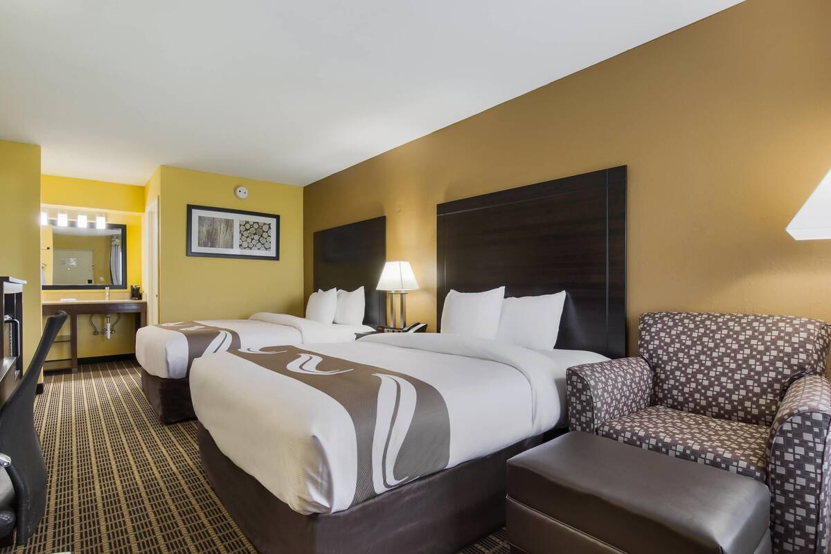 Quarto de hotel com cama grande, uma escrivaninha, uma cadeira e um banheiro visível ao fundo.