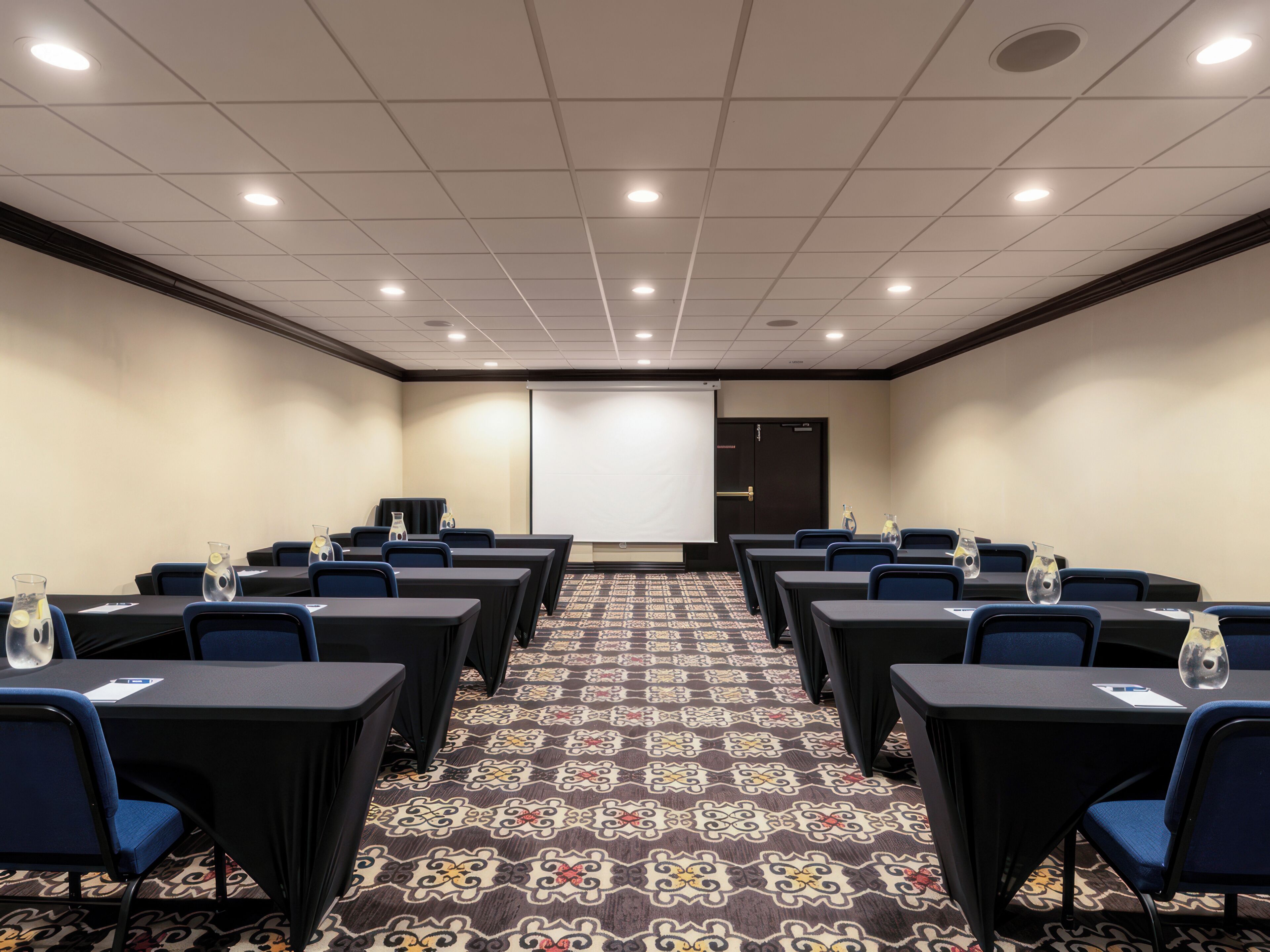 A conference room with rows of tables and chairs, a projector screen, and a patterned carpet.