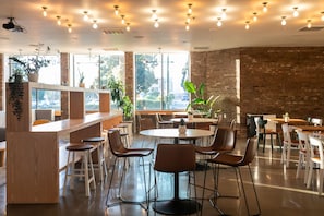 A modern cafe with round tables and chairs, large windows, and potted plants. - Hotel Burbank (Burbank)
