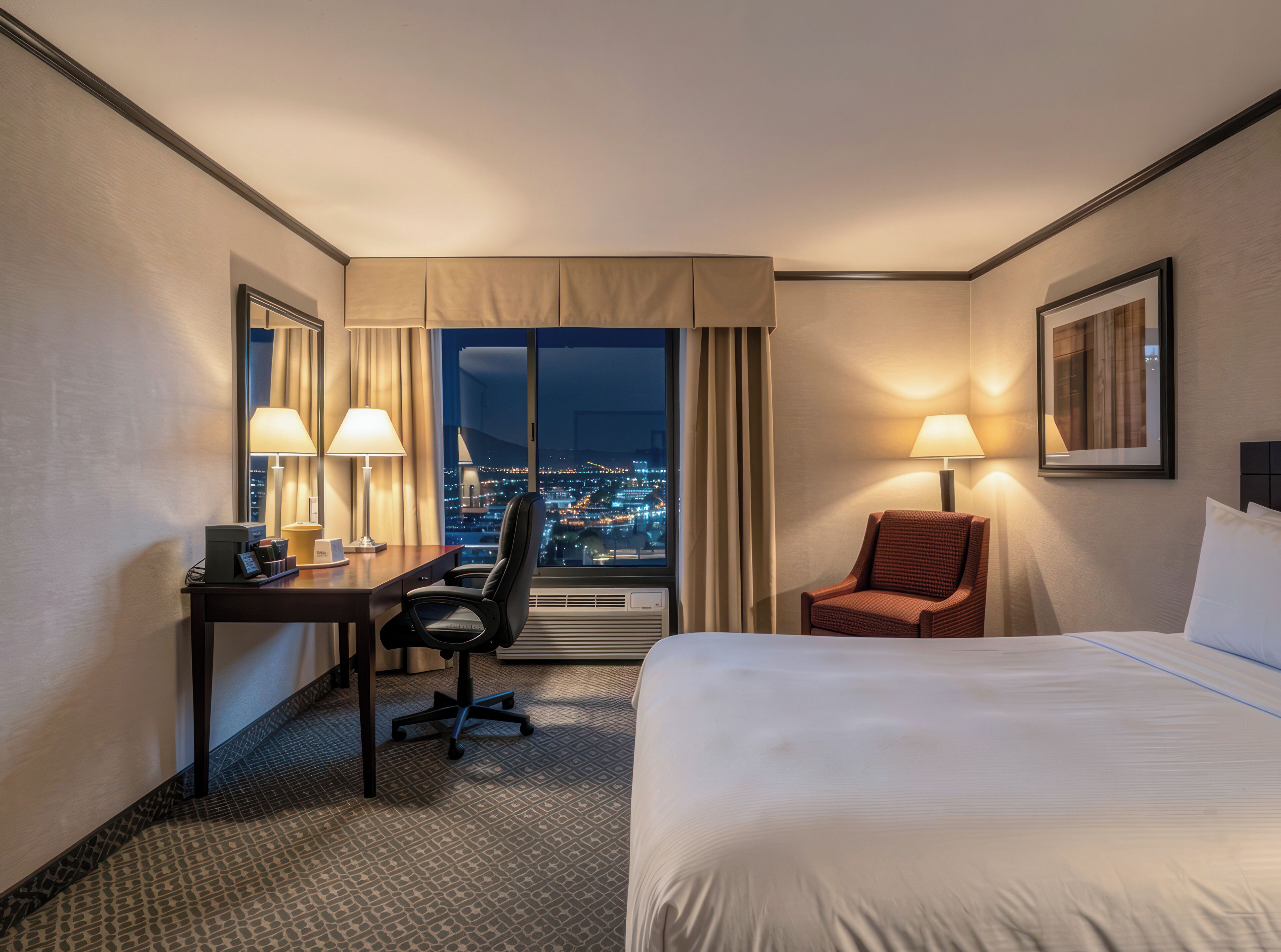 A hotel room with a bed, desk, chair, and a view of a cityscape at night.
