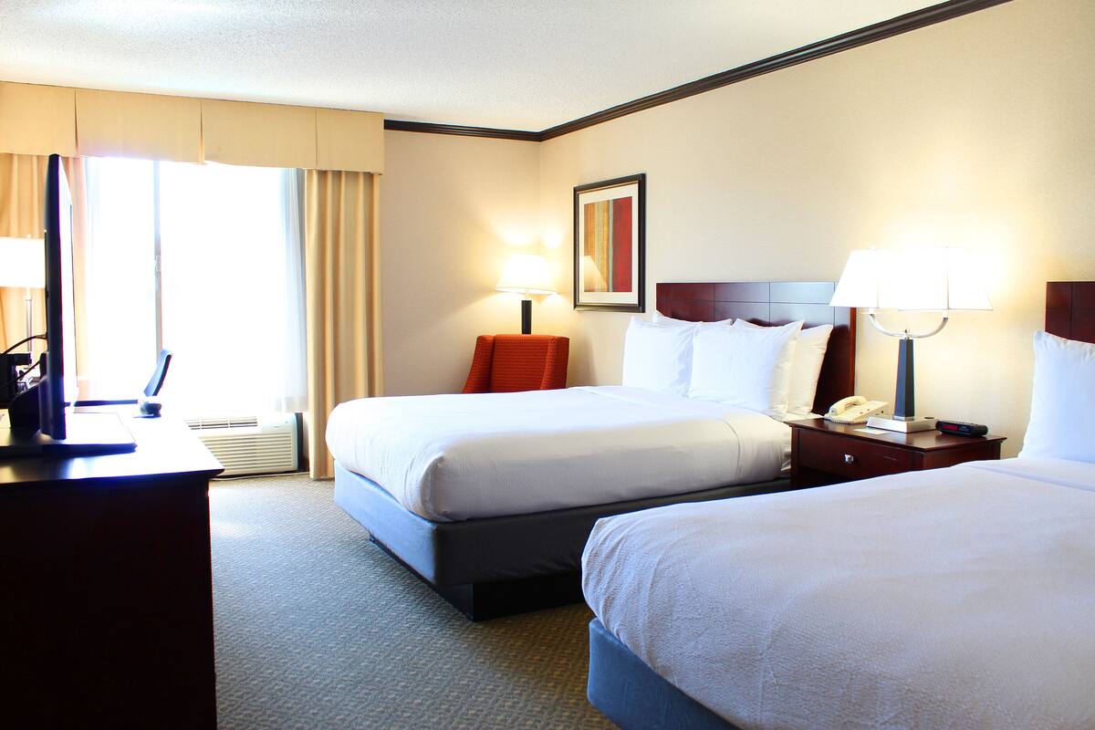 A hotel room with two beds, a desk, and a chair. There are lamps on the bedside tables and a framed picture on the wall.